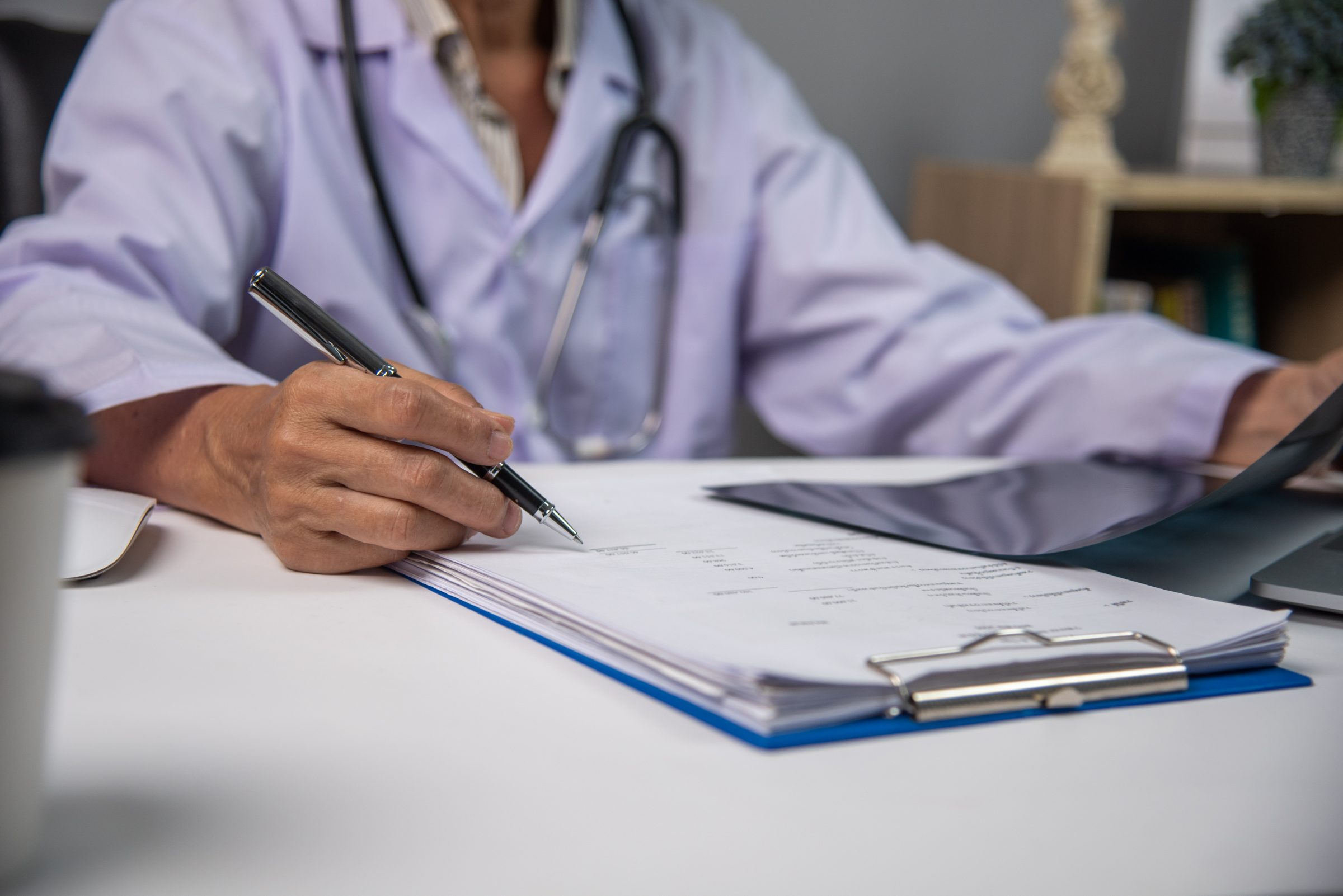 Clinician in a white coat filling in paperwork on a clipboard at a desk
